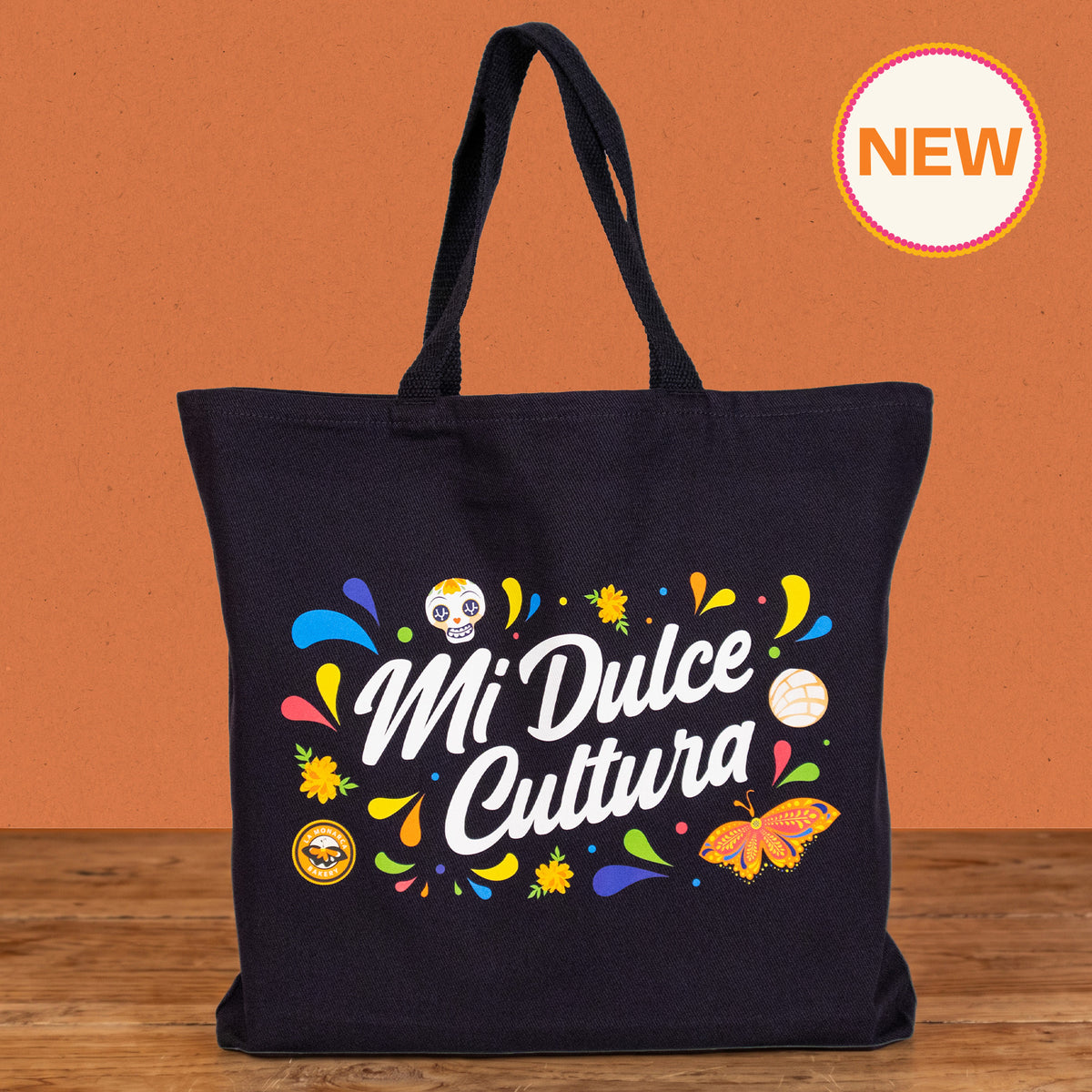 A black canvas tote bag with 'Mi Dulce Cultura' printed in white script, surrounded by colorful Dia de los Muertos-themed illustrations including sugar skulls, marigolds, a butterfly, and a cat, set against an orange background with a wooden floor and a 'NEW' badge in the top right corner.