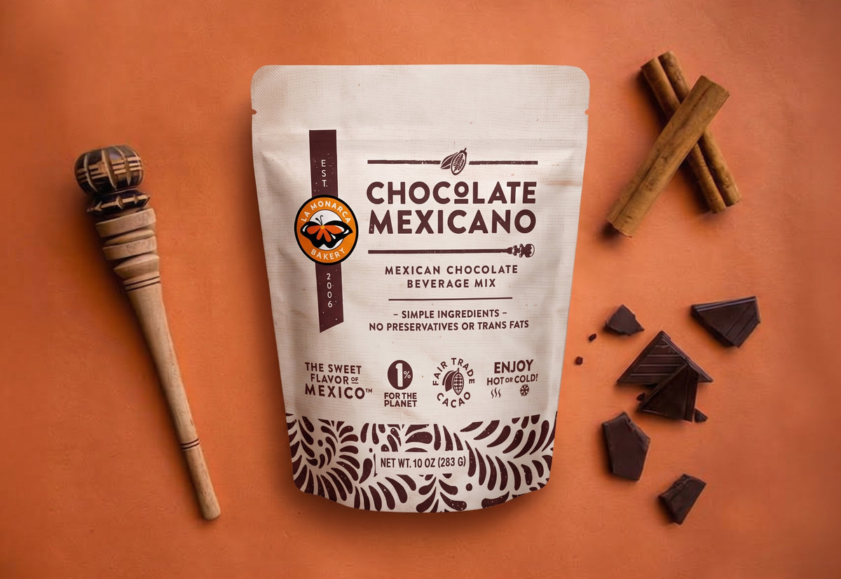 A stand-up pouch of Chocolate Mexicano Mexican Chocolate Beverage Mix is displayed on a terracotta background with a wooden spoon, cinnamon sticks, and chocolate pieces around it.
