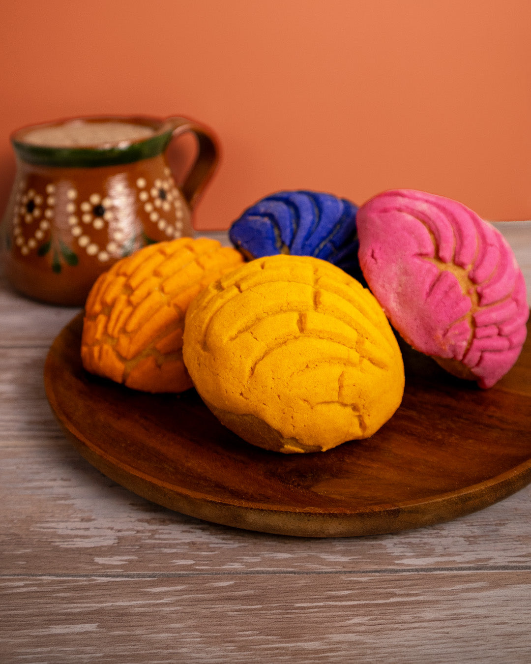 Four colorful Mexican Conchas in yellow, orange, blue, and pink, arranged on a round wooden serving platter with a ceramic mug in the background against an orange wall.